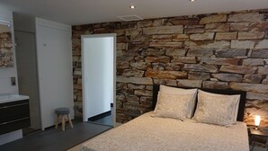 Een slaapkamer met een stenen muur, een bed met beddengoed met een patroon, een nachtkastje met een lamp, een houten kruk en een badkamer met een moderne wastafel.