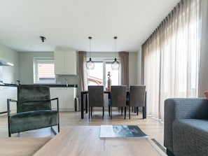 A modern kitchen with white cabinets, a dining table with chairs, and a sofa in a living area with large windows and curtains.