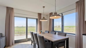 A modern dining area with a large table and chairs, large windows with curtains, and a view of a green landscape. - Vacation Home Zeeland by Oosterschelde Park (Scherpenisse)