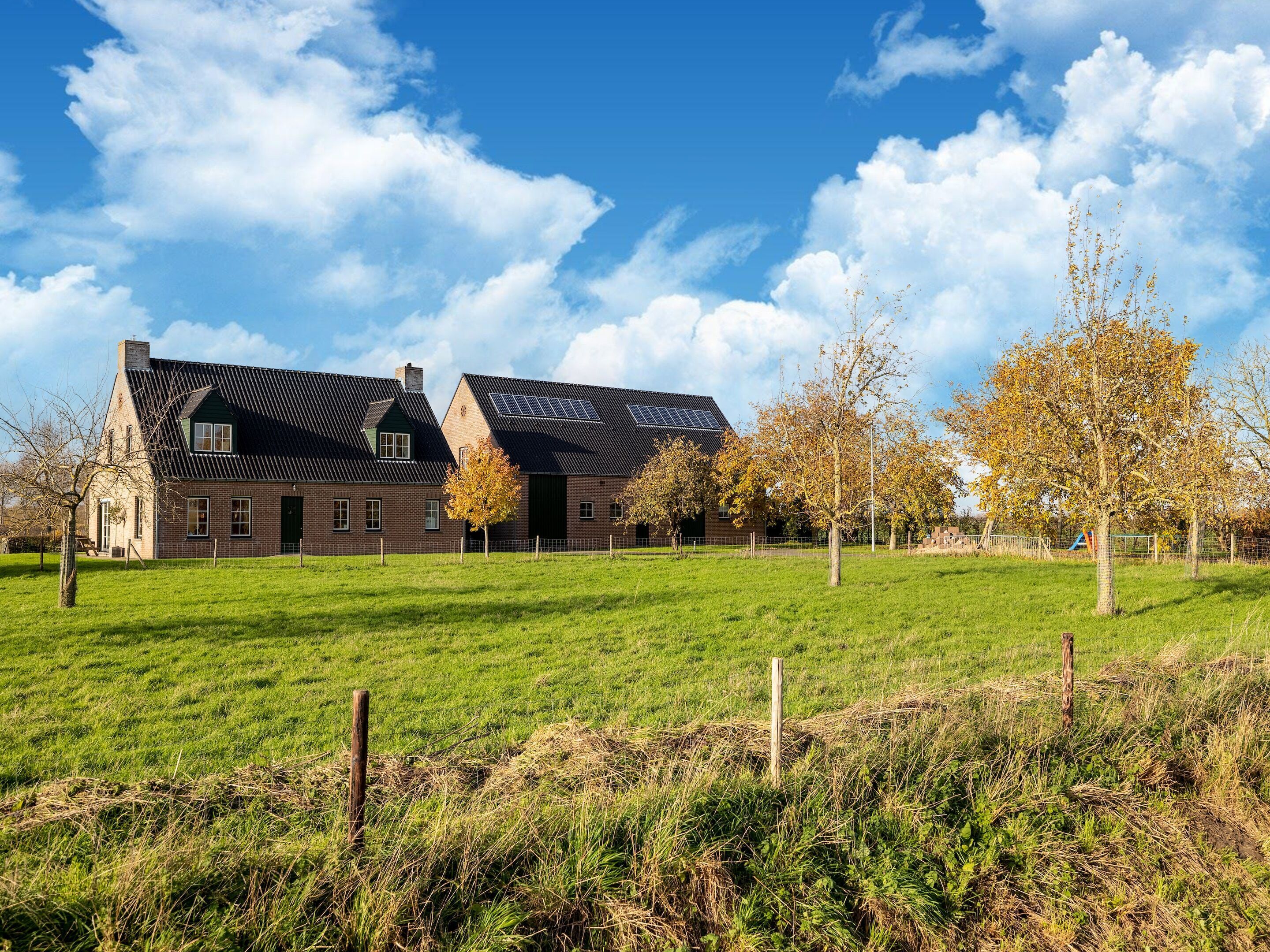 A large brick building with a sloped roof, solar panels, and a fenced green field in front.
