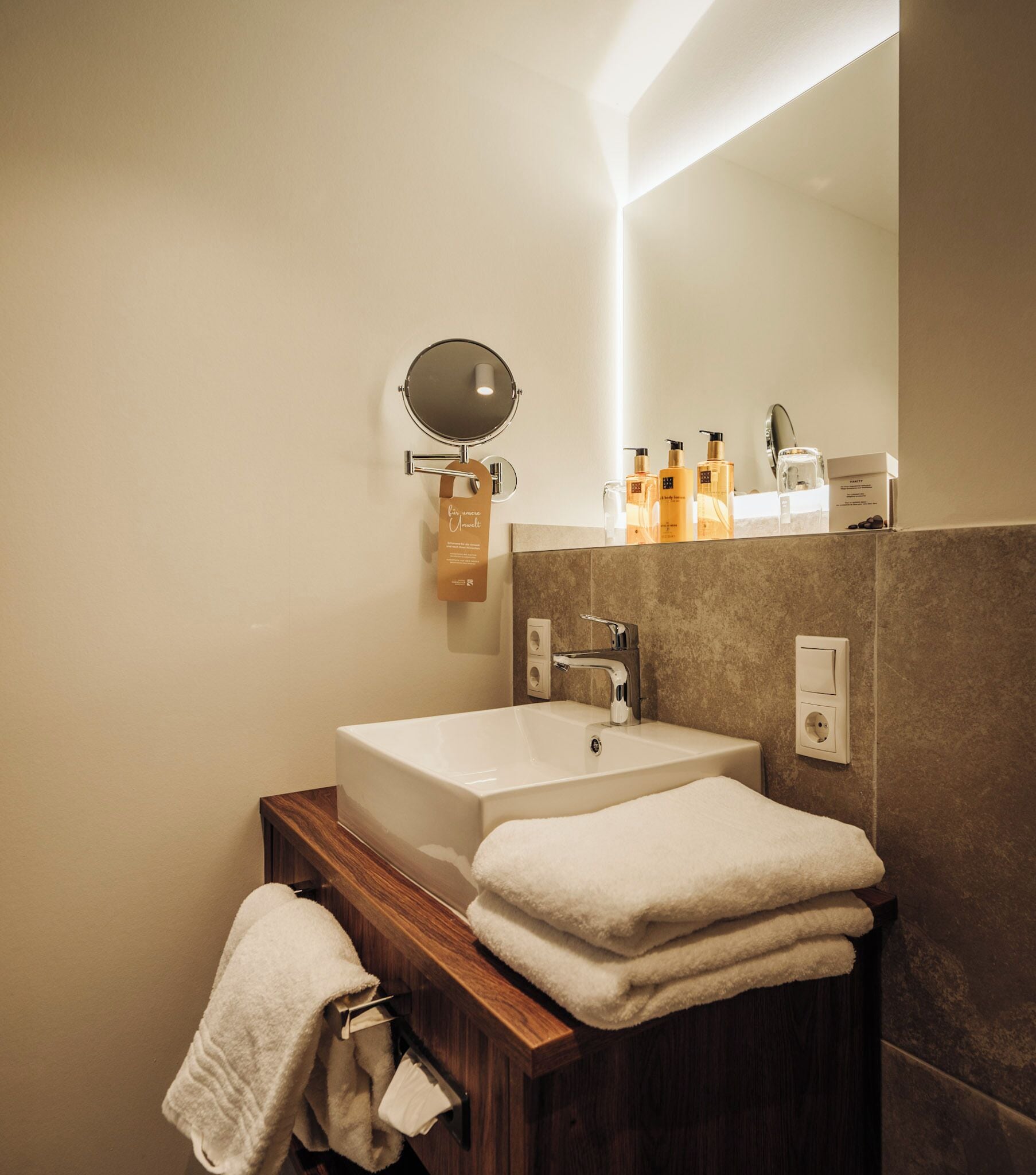 Un baño moderno con un lavamanos blanco, un espejo redondo y un mueble de tocador de madera. Hay toallas apiladas en el mueble y artículos de aseo en el lavamanos.