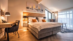 Une chambre d’hôtel moderne avec un grand lit, un bureau et une chaise. Il y a un tableau au mur et une fenêtre avec des rideaux.