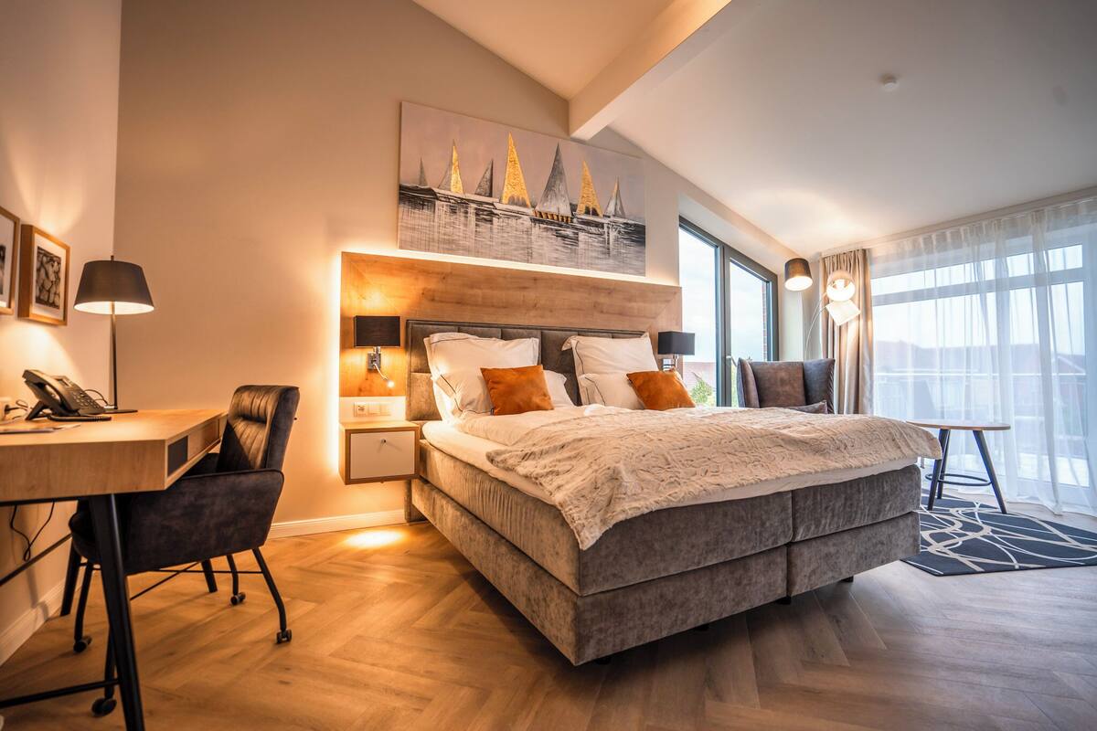 A modern hotel room with a large bed, a desk, and a chair. There is a painting on the wall and a window with curtains.
