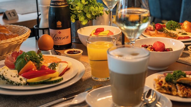 Une table à manger avec divers aliments pour le petit-déjeuner, notamment des assiettes remplies, une bouteille de champagne, des verres d’eau et de jus, et une tasse de café.