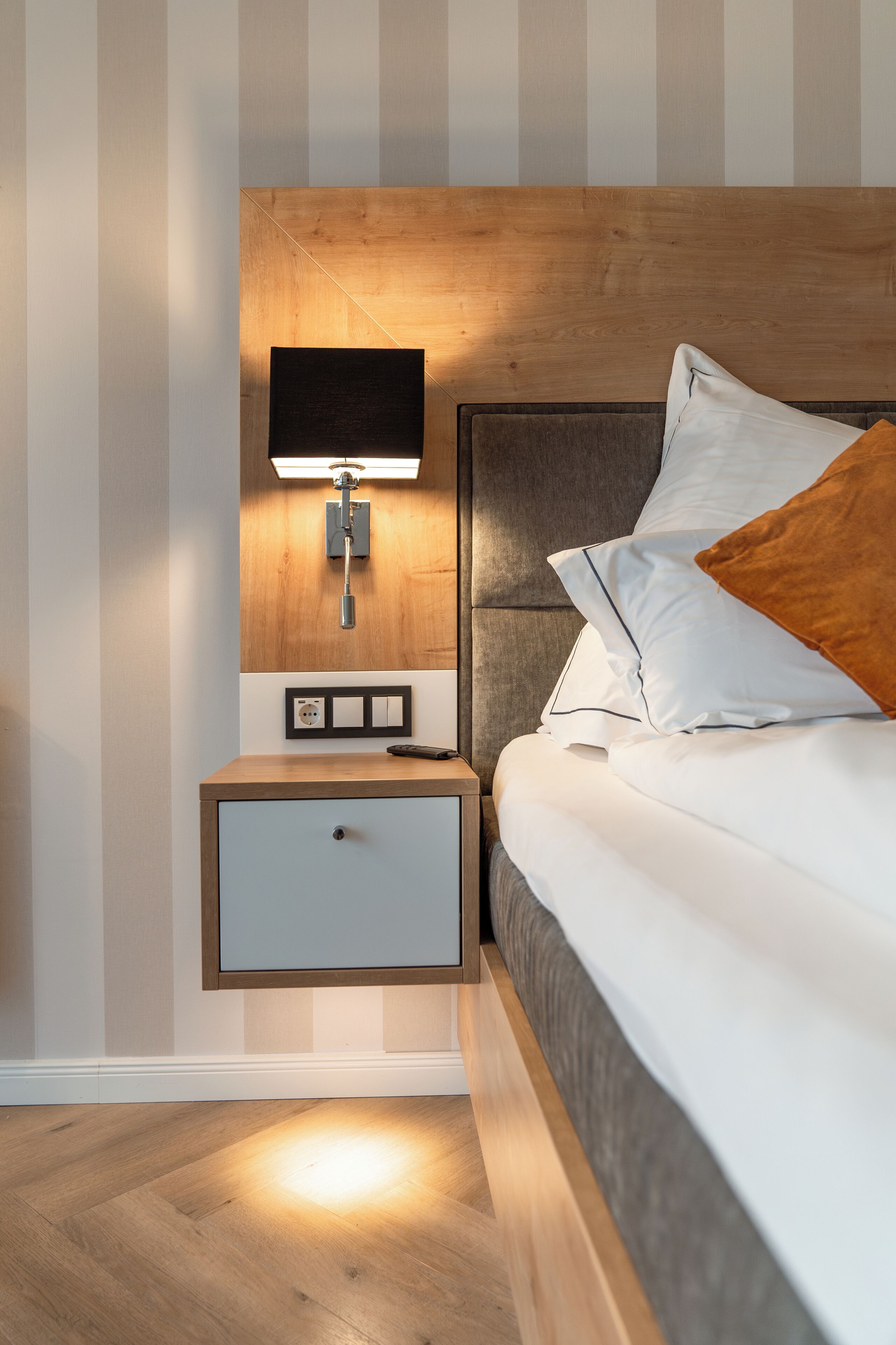 A neatly made bed with white bedding and a brown pillow, a bedside table with a lamp, and a wall-mounted light fixture.