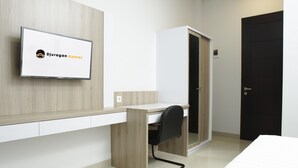 Una habitación de hotel moderna con un escritorio de madera, una silla de oficina negra, una cama y un televisor montado en la pared mostrando 'Djuran Kamar'.