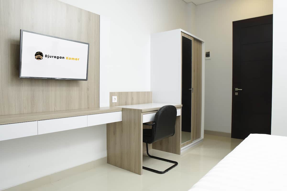 Una habitación de hotel moderna con un escritorio de madera, una silla de oficina negra, una cama y un televisor montado en la pared mostrando 'Djuran Kamar'.