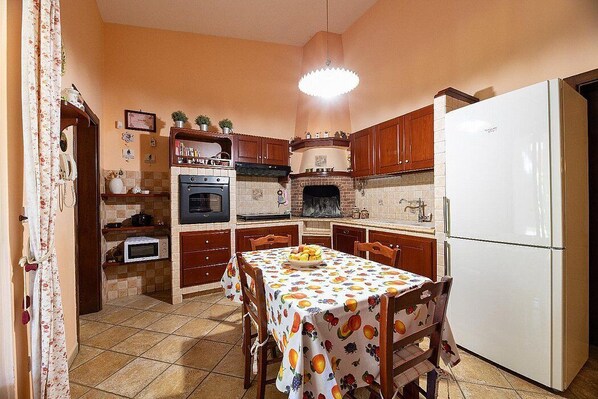 A kitchen with wooden cabinets, a white refrigerator, a dining table with a fruit-patterned tablecloth, and a built-in oven.