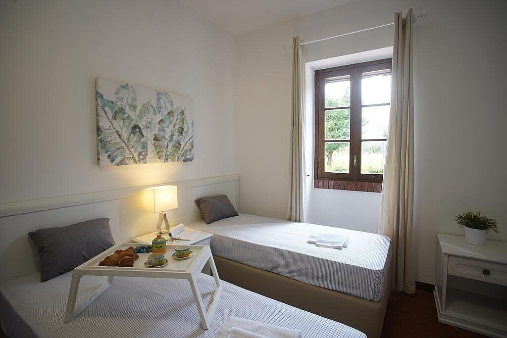 Une petite chambre propre, avec deux lits simples, une table de chevet avec une lampe, une fenêtre avec des rideaux et un tableau au mur.