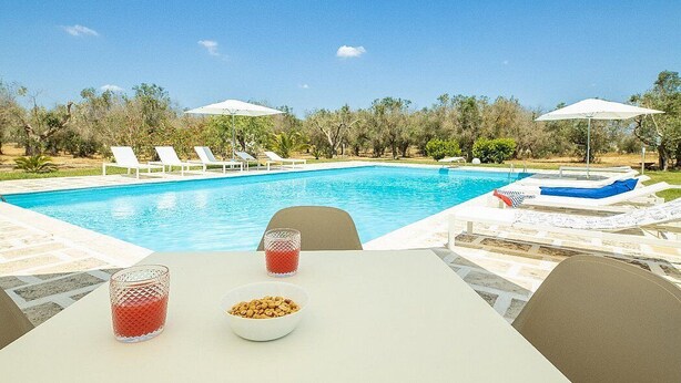 Ein Poolbereich mit Liegestühlen, Sonnenschirmen und einem Tisch mit einer Nussmischung und einem Glas rotem Getränk.
