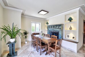 A dining area with a marble floor, wooden table, and chairs, a fireplace, and a shelf with framed pictures.