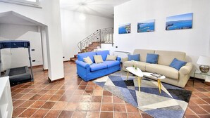 Ein modernes Wohnzimmer mit einem blauen Sofa, einem beigen Sofa und einem Couchtisch. An der Wand hängen drei gerahmte Bilder, und im Hintergrund ist eine Treppe zu sehen.