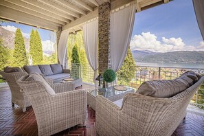 A covered outdoor seating area with wicker furniture, a glass table, and a view of a lake and mountains.