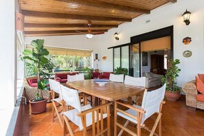 Un patio amplio con una mesa y sillas de madera, rodeado de plantas en macetas y mobiliario de mimbre.