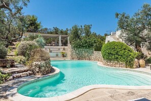 Una piscina circondata da muri in pietra e vegetazione, con una pergola e gradini che conducono a un edificio.