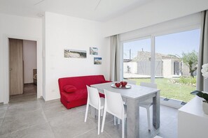 Una sala de estar moderna con un sofá rojo, sillas blancas y una mesa de comedor con cuencos rojos. Hay tres cuadros en la pared y un ventanal con vistas al exterior.