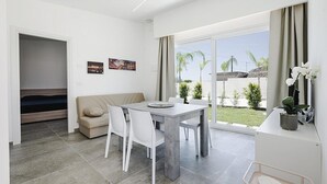 Ein modernes, helles Zimmer mit Esstisch, Stühlen, einer Couch und einem großen Fenster mit Blick auf Grünflächen und ein Gebäude.