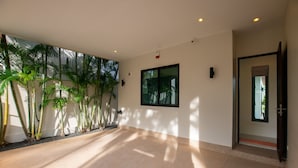 A spacious indoor area with a tiled floor, a large window, and several tall plants along the wall.