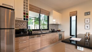A modern kitchen with a stainless steel refrigerator, wooden cabinets, a black countertop, and a window with a view of palm trees.