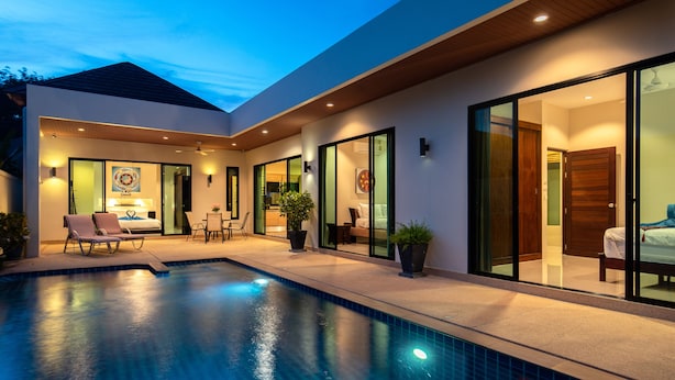 A modern house with a swimming pool, a patio with lounge chairs, and a bedroom visible through glass doors.