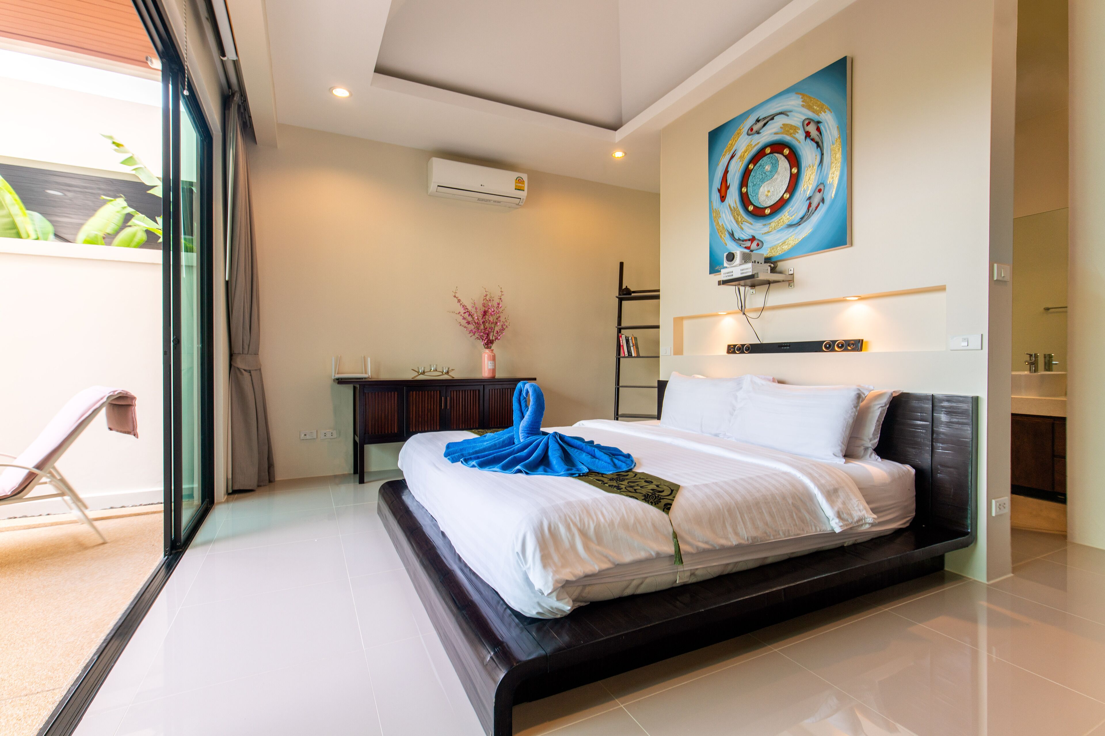 A modern hotel room with a large bed, a desk with a vase of flowers, and a large window with a view of greenery.