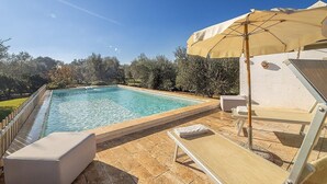 A rectangular swimming pool with clear blue water, surrounded by a tiled deck. There are lounge chairs and a sun umbrella providing shade.