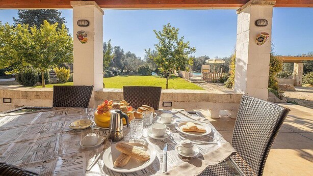 An outdoor dining area with a table set for breakfast, overlooking a garden and a building.