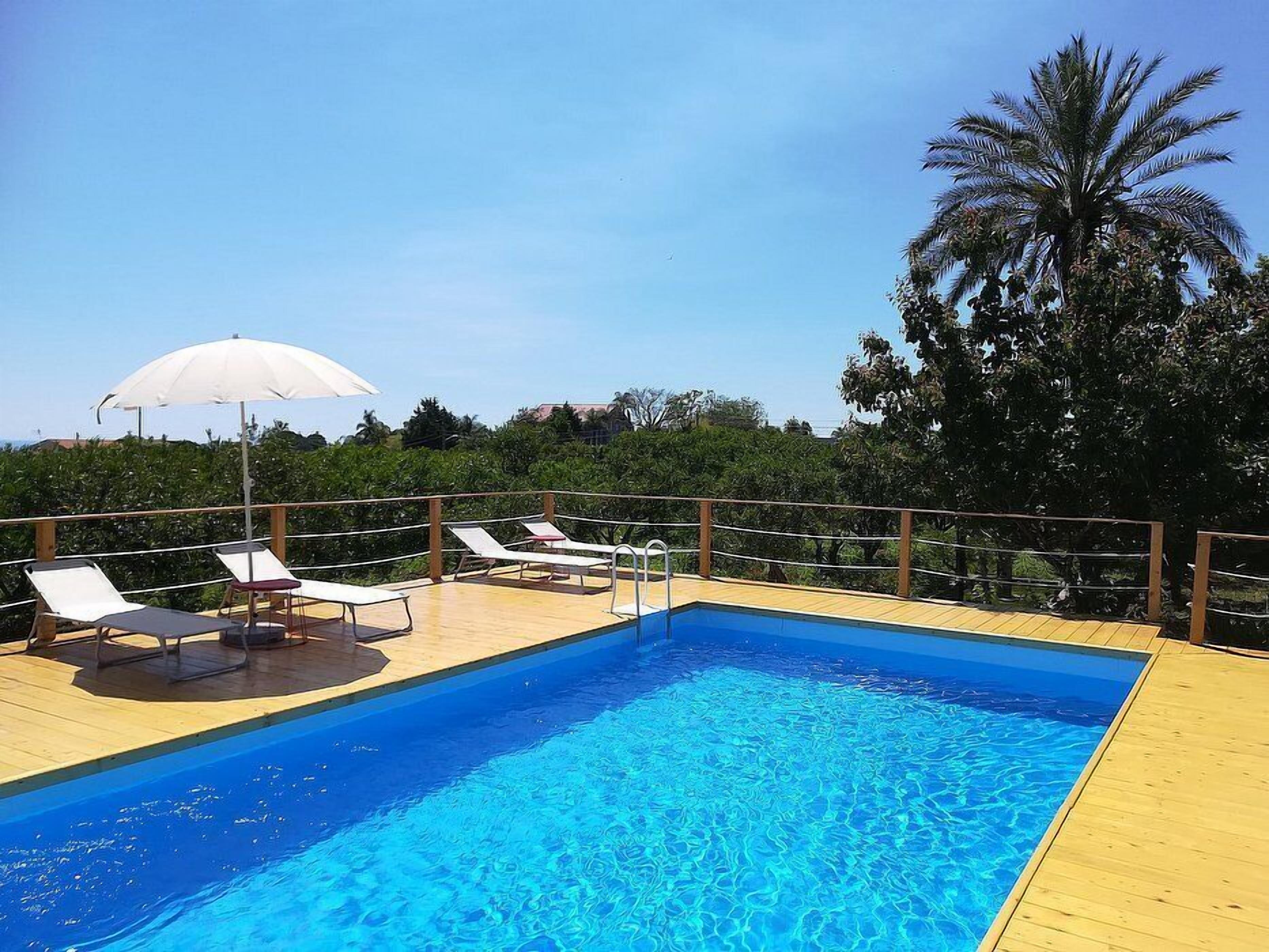 A rectangular swimming pool with clear blue water, surrounded by a wooden deck. There are white lounge chairs and a white umbrella providing shade.