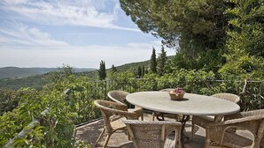 An outdoor patio with wicker furniture and a table, surrounded by greenery and overlooking a scenic landscape. - Villa Al Borgo: A welcoming two-story villa located in a characteristic village, with Free WI-FI. (Radda in Chianti)