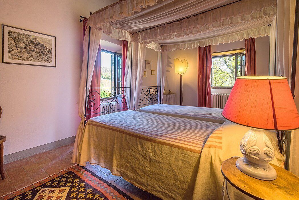 Een slaapkamer met een tweepersoonsbed, een bedlampje, een raam met gordijnen, een radiator en een lijst met een afbeelding aan de muur.