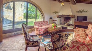 Ein gemütliches Wohnzimmer mit Kamin aus Stein, einem Fernseher und einem großen Bogenfenster mit Blick auf einen Garten.