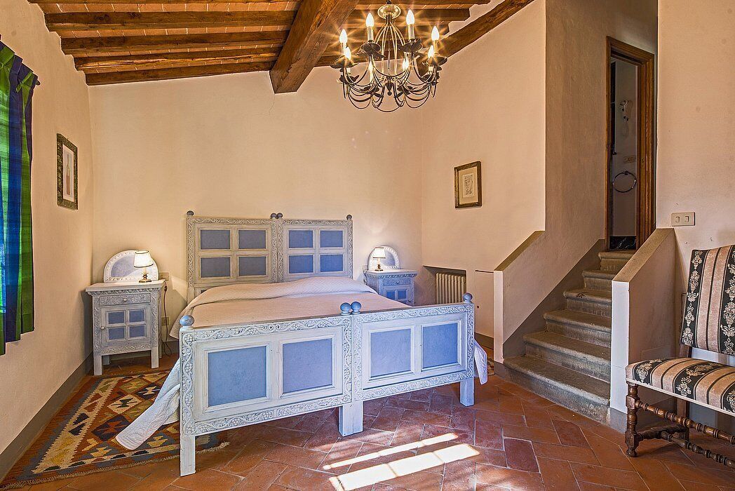 A bedroom with a large bed, two bedside tables, a chandelier, a chair, and a staircase.