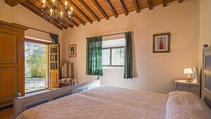 Ein Schlafzimmer mit einem großen Bett, einer Holzbalkendecke, einem Fenster mit Vorhängen, einem Nachttisch mit Lampe und einer Tür, die nach draußen führt.