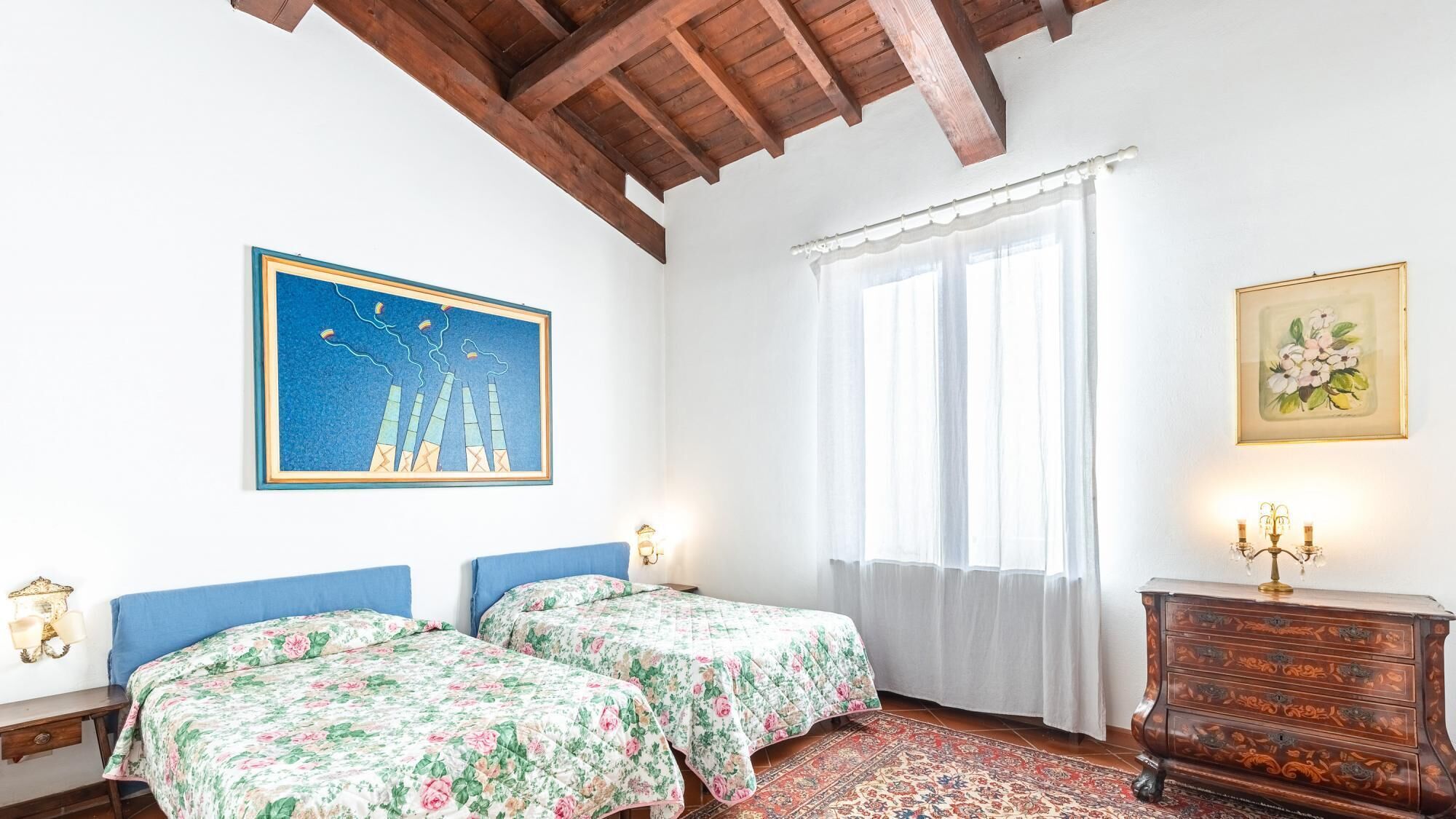A room with two beds, a wooden ceiling, a large window with sheer curtains, a wall-mounted lamp, and a framed picture.