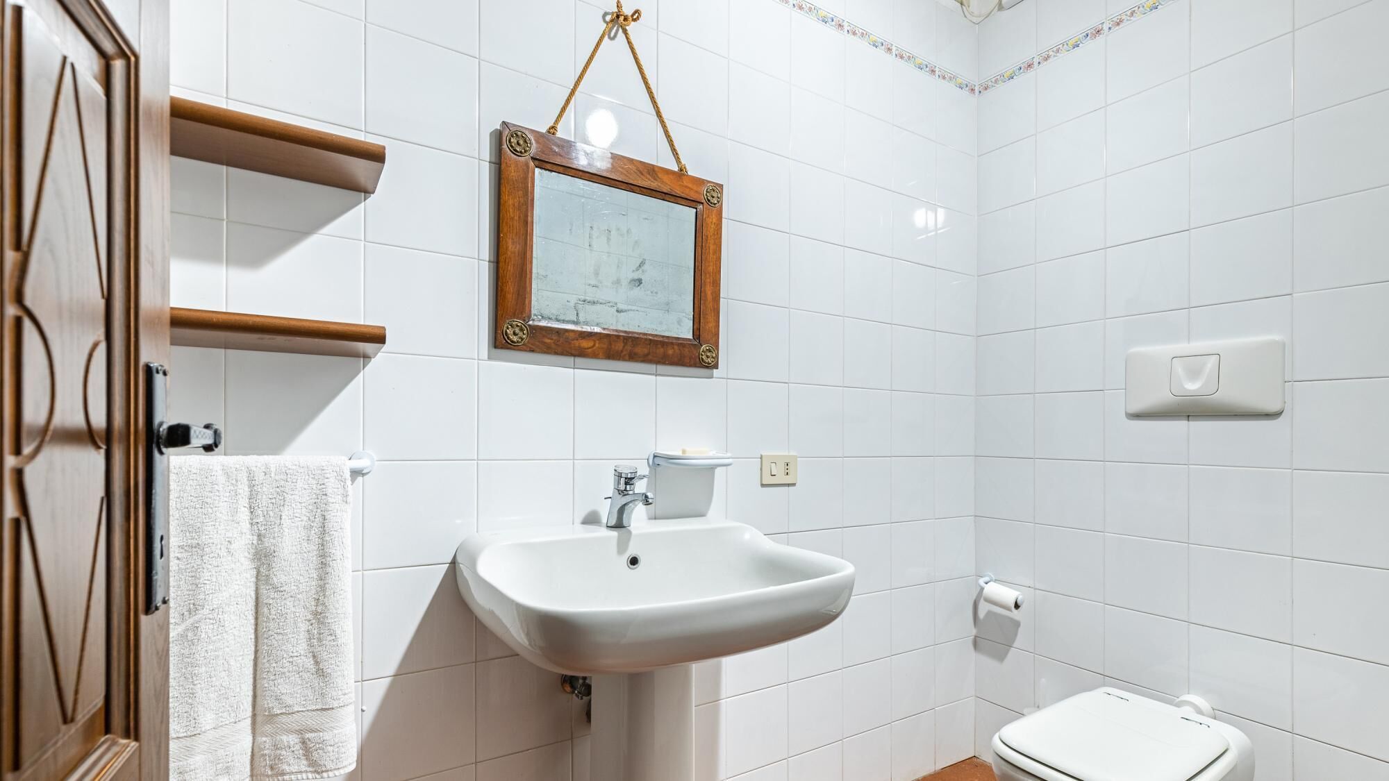 Une salle de bain avec des carreaux blancs, un miroir à cadre en bois, un lavabo blanc et des toilettes.