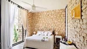 Une chambre avec un mur en pierre, un lit recouvert de draps blancs, un ventilateur de plafond et une petite table sur laquelle se trouvent un vase et des fruits.