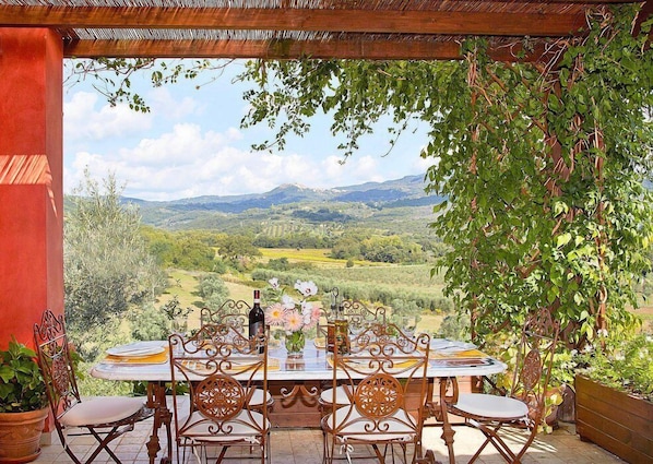 Un espace extérieur aménagé pour les repas, avec une table dressée pour six personnes, entourée de chaises, et offrant une vue panoramique sur un paysage pittoresque composé de collines et d’un ciel dégagé.