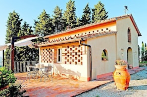 Un petit bâtiment avec un toit en tuiles, une terrasse avec des chaises et une table, et un grand pot posé sur le sol.