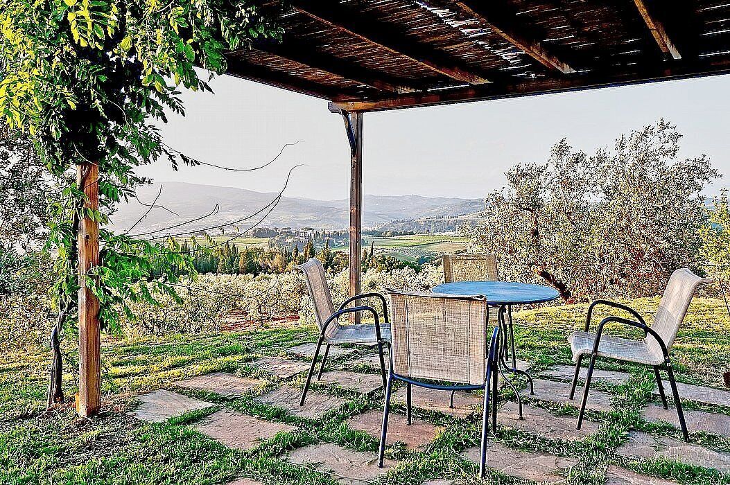 Um pátio externo com mesa e cadeiras, uma pérgula e vista para uma paisagem rural.