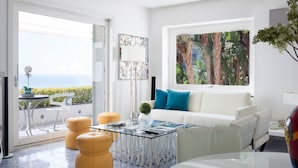 Una sala de estar moderna con una mesa de centro de vidrio, sofá blanco y un ventanal con vistas a un paisaje tropical.