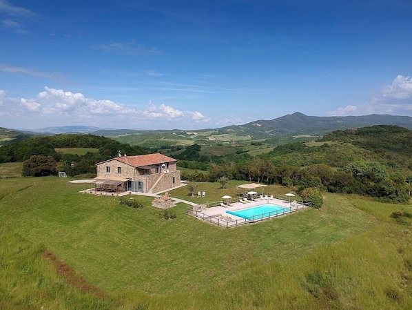 A stone house with a swimming pool, surrounded by lush green hills and a mountain in the background.