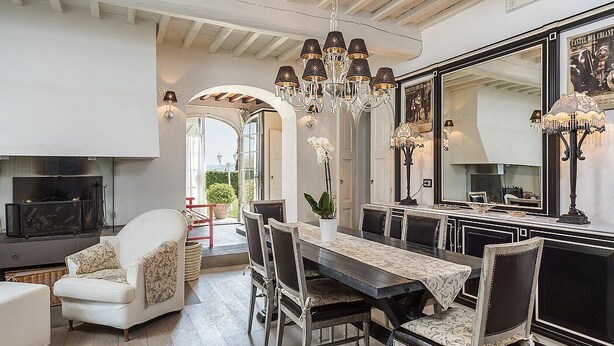 A dining area with a large table, chairs, and a chandelier. There is a fireplace and a television in the background.