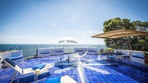 Eine Dachterrasse mit blauen Fliesen, weißen Liegestühlen und einem Tisch mit Sonnenschirmen.