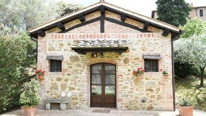 Un edificio de piedra con techo a dos aguas, una entrada central con puerta de vidrio y dos ventanas con contraventanas.
