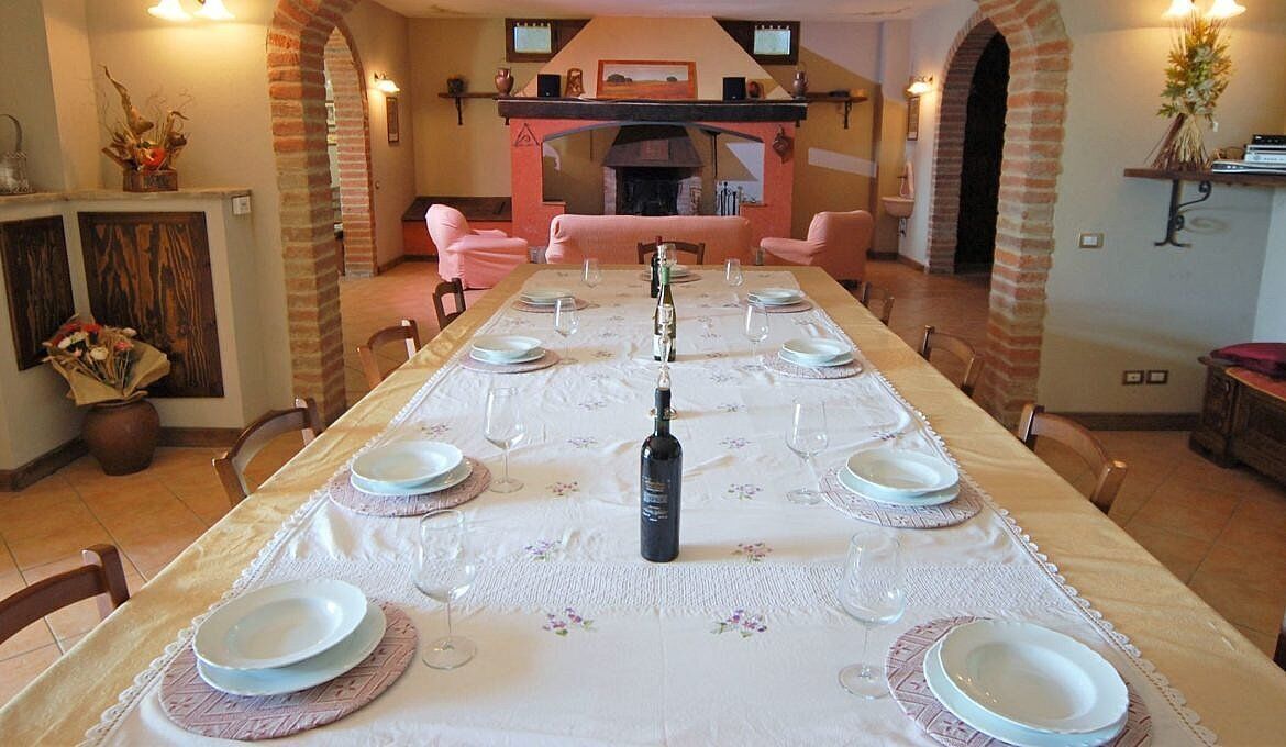 A dining room with a long table set for a meal, featuring a central wine bottle, surrounded by chairs and plates.