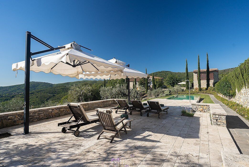 An outdoor patio with lounge chairs and umbrellas, a swimming pool, and a view of the surrounding landscape.