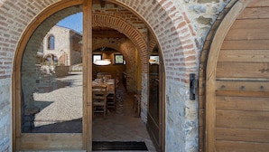 An arched doorway leading into a room with a rustic interior, featuring a wooden table and chairs, and a stone wall with a window.