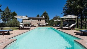 A rectangular swimming pool with lounge chairs and umbrellas on both sides, surrounded by a stone building and trees. - Villa Campanella: A characteristic and welcoming two-story age-old farm house surrounded by the greenery, with Free WI-FI. (Perugia)