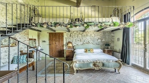 Une chambre avec un mur en pierre, un lit avec une tête de lit verte, un banc en bois, un escalier et un balcon avec une balustrade.