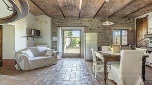 A cozy room with a stone wall, a sofa, a dining table with chairs, and a television on a shelf. - Villa Pernice: An elegant and welcoming two-story villa surrounded by the greenery, with Free WI-FI. (Castiglione D'orcia)